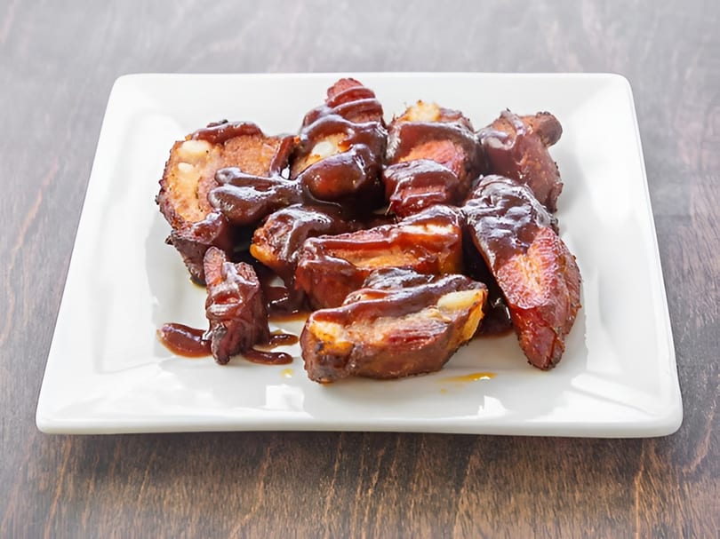 A white rectangular plate holds several pieces of saucy BBQ ribs on a dark wooden surface.