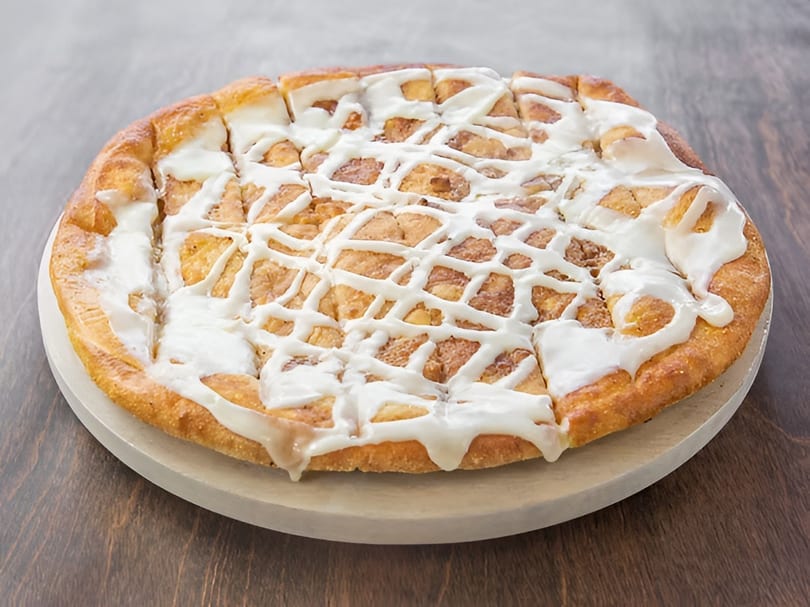 A round dessert pizza, cut into squares, topped with white icing drizzle on a wooden board.