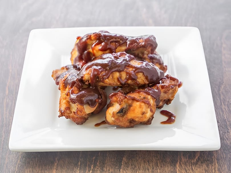 Crispy chicken wings generously coated in a rich, dark barbecue sauce on a white rectangular plate.