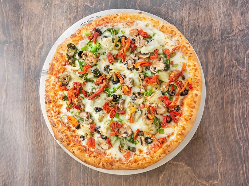 A whole vegetarian pizza with mushrooms, olives, peppers, and artichoke hearts on a wooden table.