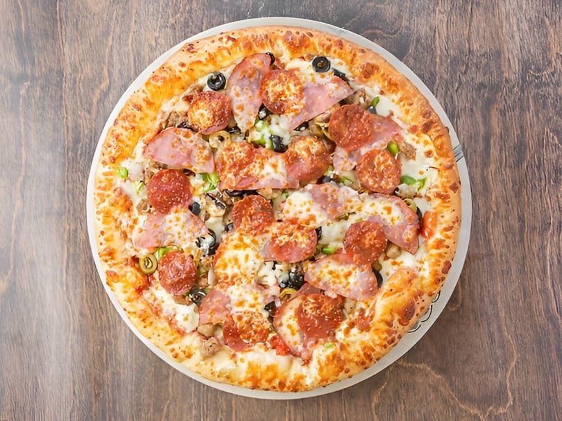 A whole pizza with pepperoni, ham, olives, and green peppers on a wooden table.